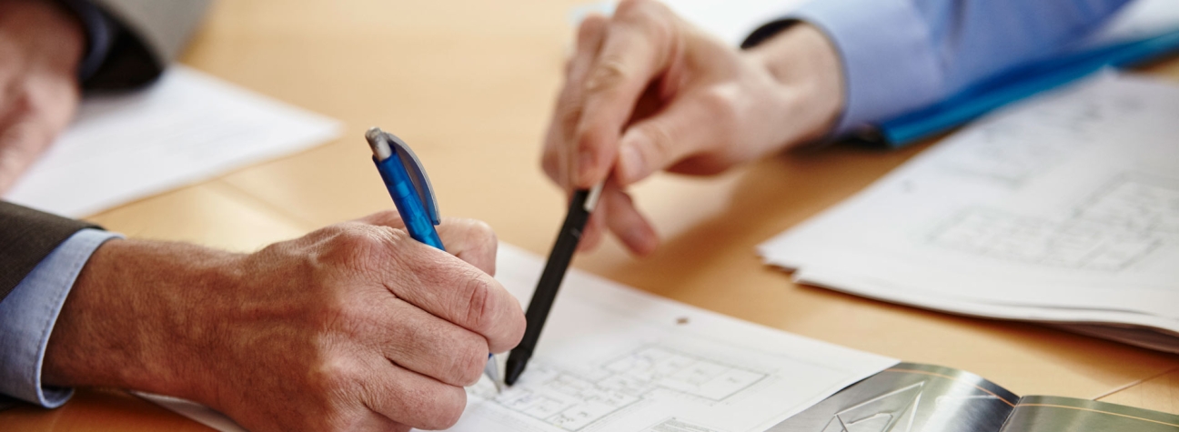 Zwei Hände mit Stift in der Hand zeigen auf ein Dokument in der Tischmitte